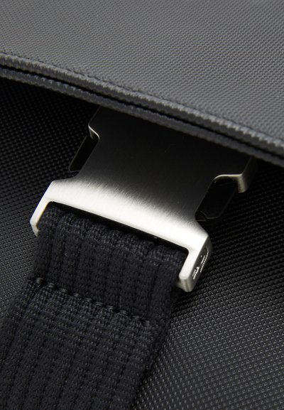 Sangle textile noire avec une boucle en métal argenté. L'arrière-plan présente une surface noire texturée, mettant en valeur les détails de conception de la sangle.