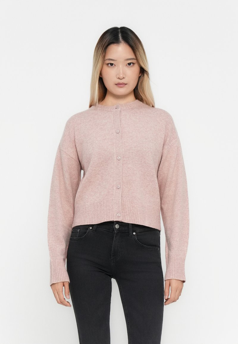 Ljust rosa stickad cardigan med rund halsringning, ribbad fåll och ärmslut, knappstängning och en avslappnad passform. I kombination med svarta jeans.