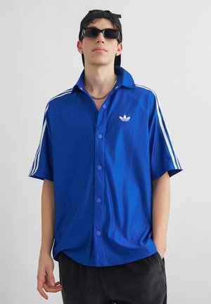 Νεαρός άνδρας που φοράει μαύρο καπέλο ανάποδα, μαύρα γυαλιά ηλίου, ασημένια αλυσίδα, μπλε πουκάμισο Adidas με λευκές ρίγες και μαύρο παντελόνι με τα χέρια στις τσέπες.