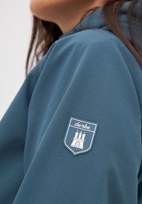 Mikina s kapucí v modrošedé tkanině. Vybavena výraznou nášivkou se štítem a designem hradu na levém rukávu. Hladká textura.
