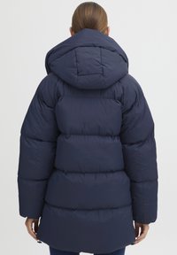 Navy puffer jacket with a hood, quilted texture, and a slightly cinched waist. Features a long, oversized design with elastic cuffs.