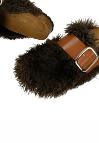 Hnědé chlupaté pantofle s plyšovým textilním svrškem, hnědým koženým páskem se stříbrnou přezkou a texturovanou podrážkou pro lepší přilnavost.