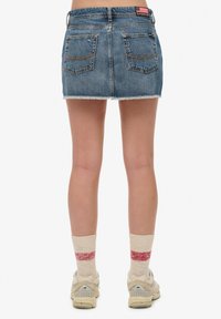 Jupe en jean mini de couleur bleu clair avec un ourlet effiloché, dotée de deux poches arrière et de coutures contrastées. Portée avec des chaussettes et des baskets.