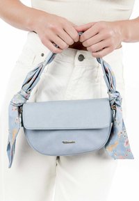 Helleblaue Lederhandtasche mit einer geschwungenen Form, ausgestattet mit einer blumengemusterten Stoffband, das am Griff befestigt ist, und einem Metallemblem.
