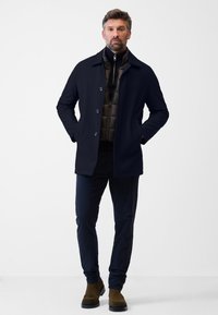 Marinblå kappa med knapprad, lagd över en mörk vadderad väst. Parat med mörka byxor och bruna mocka ankelskor. Enkel, stilren design.