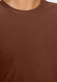 Brun crew neck t-shirt med ett texturerat mönster av Nike-logotyper och ordet "AIR" i en upprepande design. Slät tygfinish.