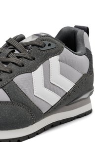 Szary but sportowy z teksturowaną cholewką z zamszu i siateczki. Posiada białe akcenty w kształcie szewronów, brązowe sznurówki oraz stabilizujący zapiętek.
