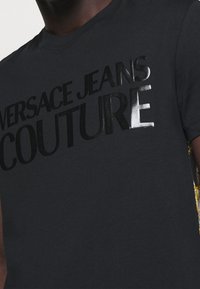 Schwarzes T-Shirt mit glänzend schwarzem "VERSACE JEANS COUTURE"-Schriftzug, getragen von einer dunkelhäutigen Person, die den oberen Brustbereich und kurze Ärmel zeigt.