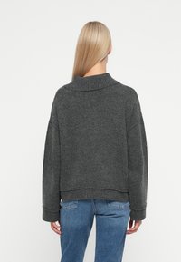 Mørkegrå strikkegenser med ribbete krage, senket skuldre og en kort hem. Brukt sammen med lyseblå jeans som har en avslappet passform.