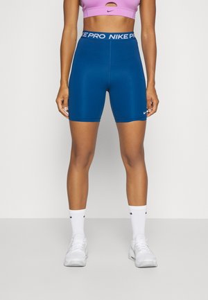 Kobieta ubrana w niebieskie, wysokie szorty rowerowe Nike Pro, fioletowy stanik sportowy, białe skarpety Nike oraz białe buty sportowe, stojąca na jednolitym tle.