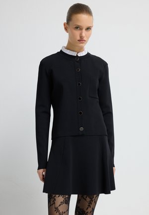 Veste noire courte avec six boutons sur le devant, dotée d'une petite poche. Associée à une jupe noire évasée et des collants à motifs.