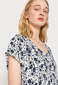 Top com estampa floral em branco com flores azul-escuras. Apresenta decote em V e mangas curtas enroladas. Tecido leve com uma textura suave.