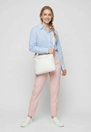 Junge Frau mit blondem Zopf trägt ein blau kariertes Hemd, rosa Hose, weiße Sneakers und trägt eine weiße Umhängetasche.