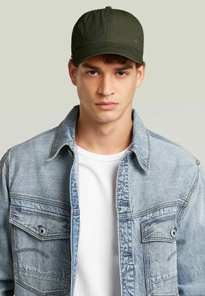 Jonge man met een donkergroene pet, lichtblauwe denim jas over een wit T-shirt, kijkt recht vooruit met een neutrale uitdrukking.