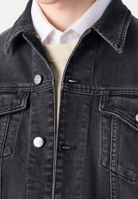 Svart denimjacka med klassisk krage, framfickor, silverdetaljer och subtil textur, lager på lager över en vit skjorta och tröja.