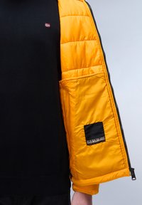 Schwarzer Pullover unter einer offenen orangen Steppjacke. Die Jacke hat eine schwarze Tasche mit einem Logo und einem gesteppten Design, das einen strukturellen Kontrast schafft.
