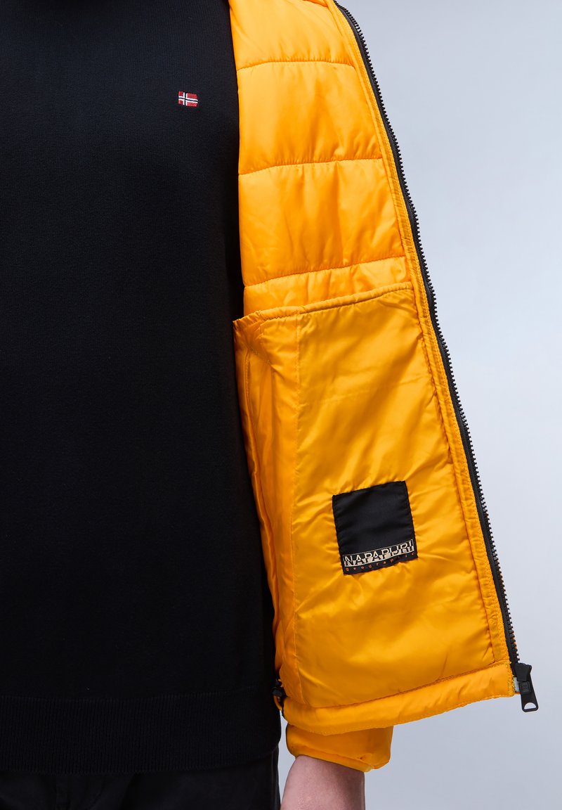 Maglione nero sotto un giubbotto arancione imbottito aperto. L'imbottito ha una tasca nera con un logo e un design trapuntato, creando un contrasto tattile.