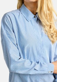 Mėlynos ir baltos spalvų dryžuotas ilgomis rankovėmis marškinėlis su sagotu apykakle, papuoštas mažais dekoratyviniais karoliukais ir laisvo silueto dizainu.