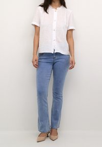 Camicetta bianca in lino a maniche corte con colletto arricciato, abbinata a jeans dritti azzurri e décolleté beige a punta.