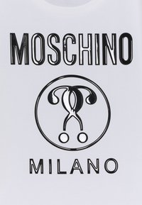 Camiseta blanca con el texto en negrita negro "MOSCHINO" y "MILANO", con un logotipo gráfico de tijeras dentro de un círculo. Textura de superficie suave.