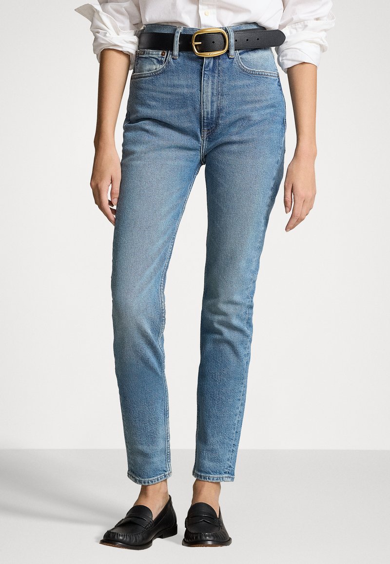 Blue high-waisted jeans with a slim fit, featuring a brown leather belt with a large gold buckle and black loafers. Smooth denim texture.