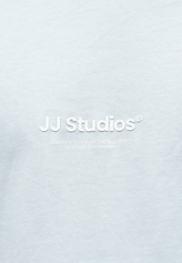 Jasnoniebieska koszulka z białym nadrukiem logo "JJ Studios" oraz tekstem "Brought to you by THE J.J. CREW™ Designed in Denmark." Gładka faktura.