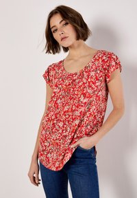 Blusa floral roja con mangas cortas, escote redondeado y diseño fluido. Presenta una mezcla de patrones florales en blanco y verde sobre una tela ligera.