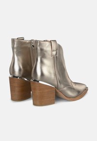 Botines de tobillo metálico dorado con punta curva, cremallera lateral y costura decorativa. Presentan un tacón de madera grueso con un acento plateado.