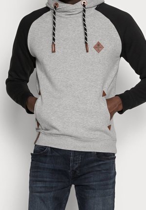 Man draagt een grijze hoodie met zwarte mouwen en gestreepte trekkoorden, handen in de zijzakken, gecombineerd met donkerblauwe spijkerbroek.