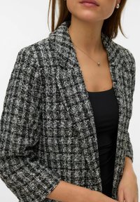 Blazer in tweed bianco e nero con finitura testurizzata, rever a incavo e maniche a tre quarti. Indossato sopra una maglia nera.