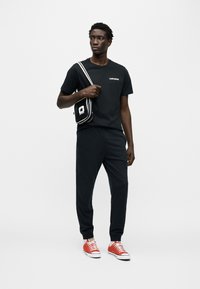 Μαύρο βαμβακερό μπλουζάκι με λευκό λογότυπο, μαύρη φόρμα, και κόκκινα πάνινα παπούτσια. Μικρή μαύρη τσάντα ώμου με λευκό λογότυπο αστεριού.