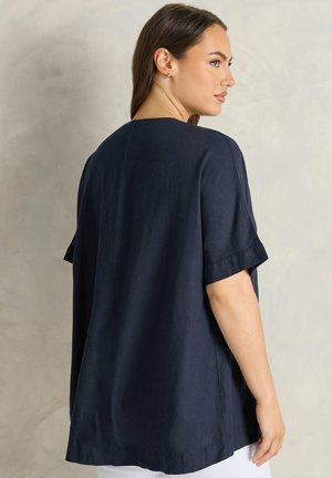 Mujer de cabello oscuro que lleva una blusa azul marino de manga corta y holgada, posando de perfil sobre un fondo neutro.