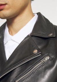 Černá kožená motocyklová bunda s výrazným zipem, stříbrnými kovovými detaily a hladkou texturou. Pod ní bílá košile s límečkem.