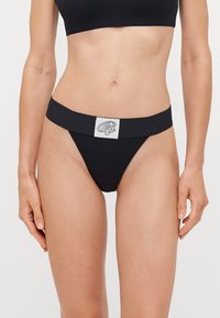 Black thong underwear with a wide waistband featuring a logo patch. Made of smooth fabric, it has a minimalist design and triangular shape.