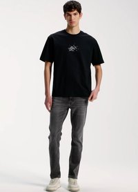 T-shirt de algodão preta com estampa gráfica branca no peito, combinada com jeans cinzentos e ténis creme. Design simples e casual.