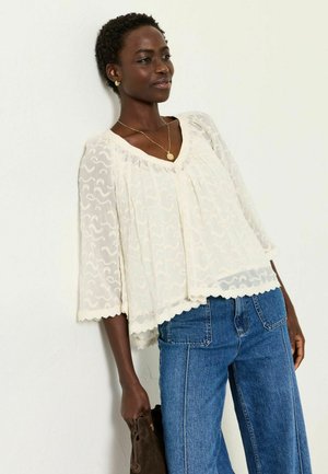 Woman wearing a white lace blouse with scalloped hem, blue jeans, gold necklace, and earrings, holding a brown suede bag against a white wall.