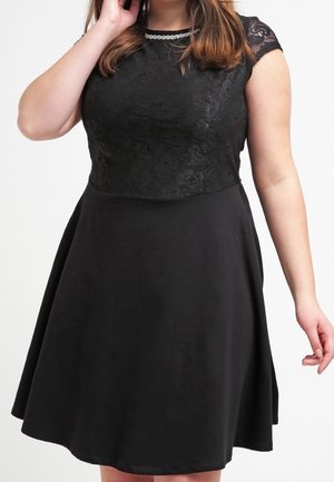 Kobieta w czarnej sukience z koronkowymi, kapturzastymi rękawami i koronkowym kwiatowym gorsetem, ozdobiona perłowym naszyjnikiem, pozująca na białym tle.