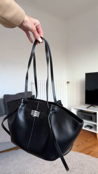 Fekete bőrszatyor két hosszú pánttal, ezüst kapoccsal, és ívelt sziluettel. Simafejű anyag minimális díszítéssel.