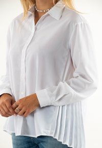 Blusa blanca de botones con mangas largas, que presenta un diseño plisado en un lado y una textura suave, combinada con jeans de color azul claro.
