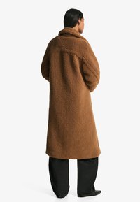 Rjav plašč iz flisa s širokim ovratnikom, teksturirano tkanino in sproščenim krojem, kombiniran s črnimi širokimi hlačami.