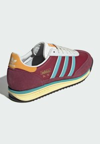 Tenisky Adidas SL '72 v tmavě červené semišové kůži s světle modrými pruhy, bílými koženými detaily a žlutou podrážkou. Obsahují polstrovaný límec.