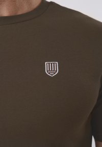 Κοντινό πλάνο ενός ατόμου που φοράει σκούρο πράσινο μπλουζάκι με μικρό λευκό έμβλημα ασπίδας με κάθετες ρίγες στο στήθος.