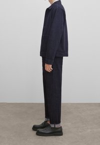 Tummansininen farkkutakki kauluksella, etutaskuilla ja siihen sopivat housut. Mustat kengät kuvioidulla pohjalla, housujen lahkeet käärityt.