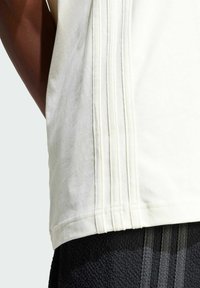 Light cream sleeveless top with side pleats, made of soft fabric. The hemline is curved, paired with textured black athletic shorts.