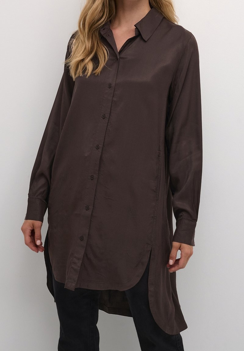 Femme portant une robe-chemise à manches longues marron foncé avec col et fentes latérales, associée à un pantalon noir.