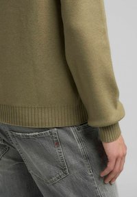 Maglione color khaki con polsini e orlo a costine, abbinato a jeans grigi. Il tessuto appare morbido e lavorato a maglia, con una finitura testurizzata.