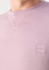Gros plan d'une personne portant un sweat-shirt à col rond rose clair avec un petit logo carré "BOSS" sur la poitrine.