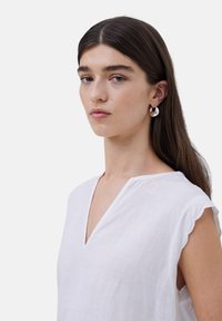 Top de lino blanco con escote en V, mangas cortas con ondas y un corte holgado. La modelo lleva pendientes hoop metálicos. Textura sencilla.