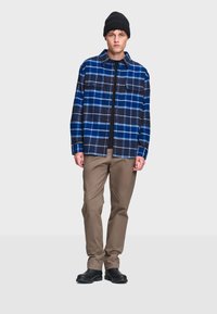 Sininen ja musta ruudullinen flanellipaita, jossa on kaksi rintataskua, harmaa t-paita alla, khaki-housut ja musta kengät. Yksinkertainen, rakennettu muotoilu.