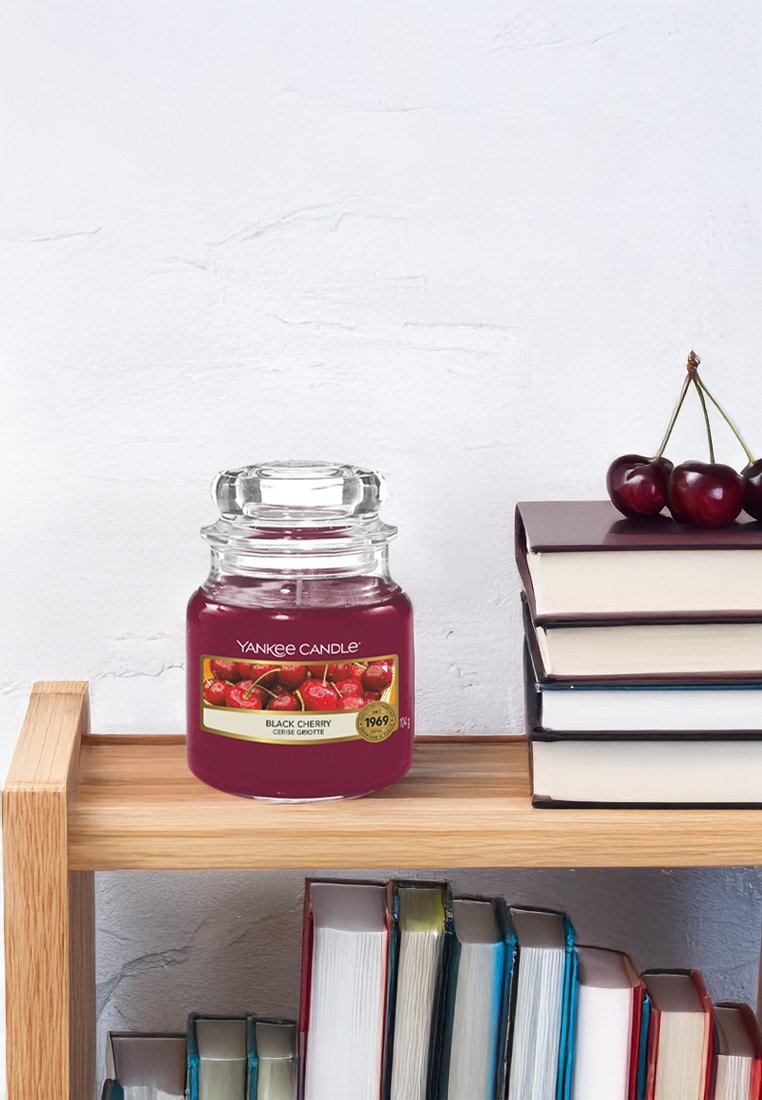Vela Yankee Candle en un tarro de vidrio transparente, llena de cera burdeos oscuro, etiquetada como "Cereza Negra", sobre una estantería de madera al lado de libros apilados.